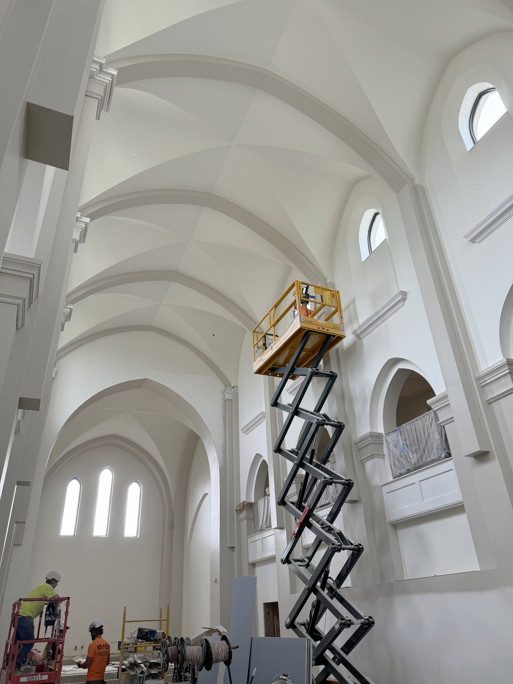 A finisher works from a scissor lift beneath a vaulted ceiling while two crew members work below.